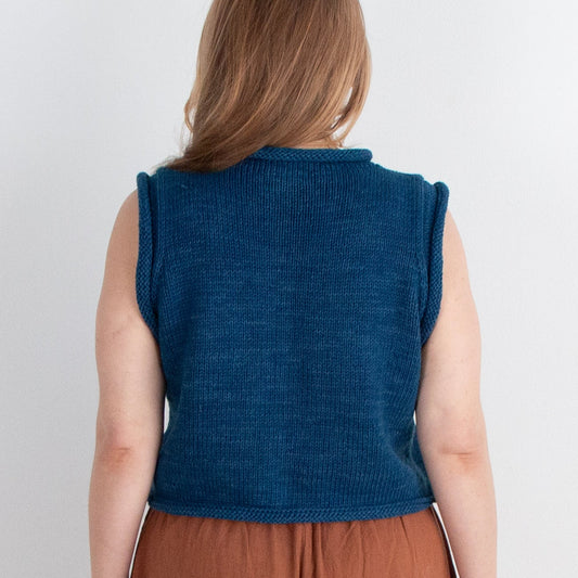 Woman wearing a blue sleeveless top and brown pants standing against a white wall.