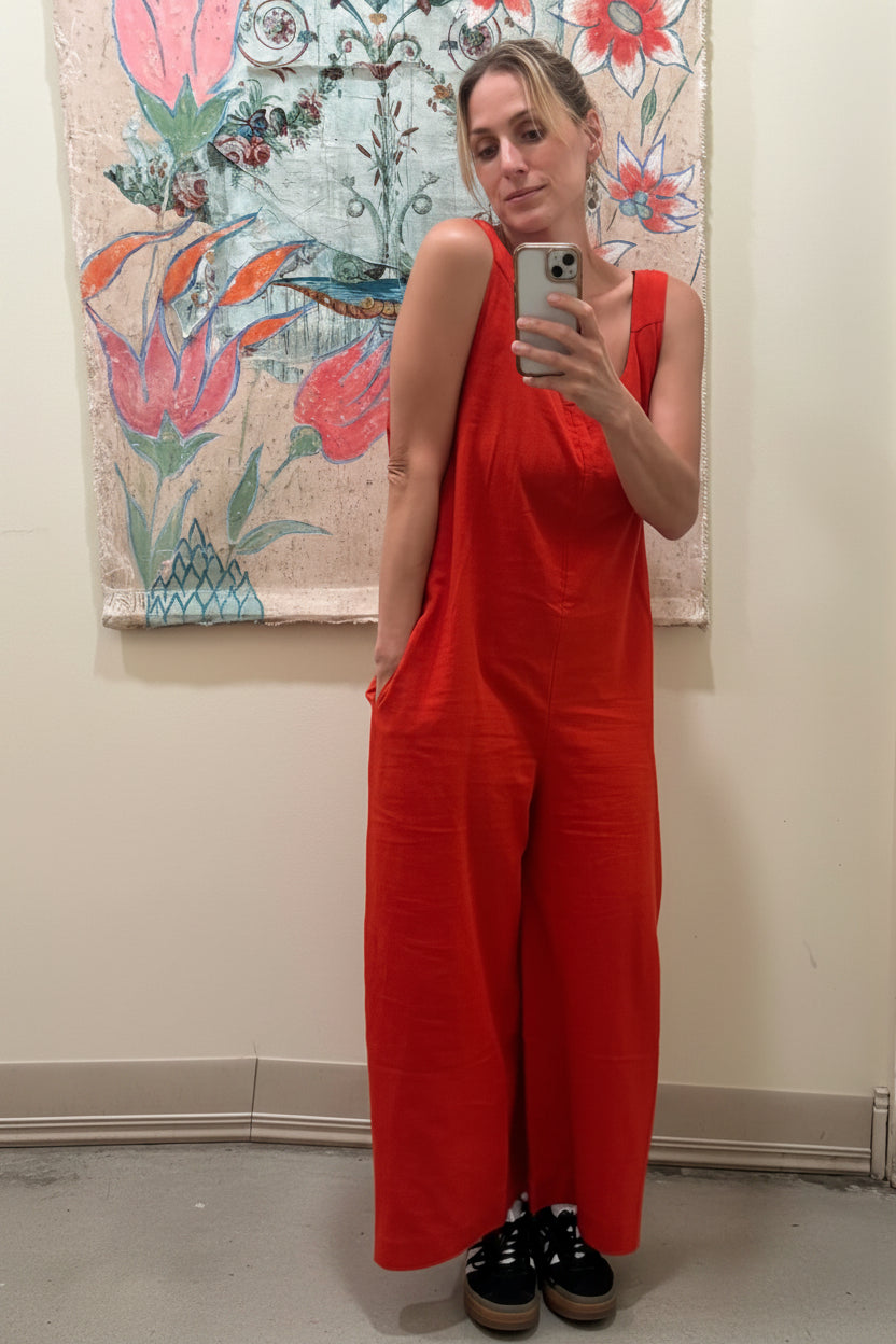 Person wearing a red jumpsuit taking a mirror selfie in front of a floral tapestry.