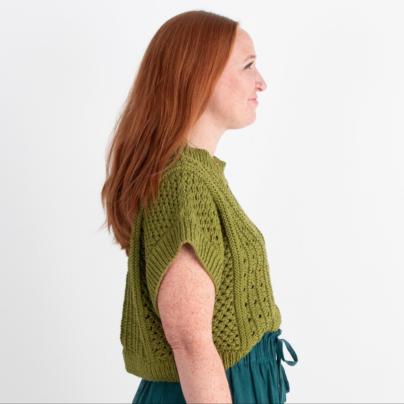 Woman wearing a green knitted top and teal pants against a white background